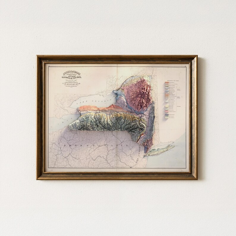 May include: Framed vintage map of New York State with a 3D topographic relief. The map features a detailed depiction of the state's terrain, with varying colors indicating elevation. The frame is a warm, dark brown color.