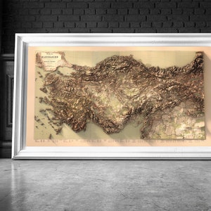 May include: A vintage, detailed relief map of Asia Minor, also known as Anatolia, with the title "Kleinasien" in German. The map is framed in a white frame and displayed against a black brick wall.