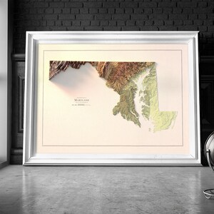 May include: A framed 3D relief map of Maryland with a beige background. The map is in shades of brown, green, and yellow, showing the state's topography. The word "Maryland" is printed in black at the bottom of the map.