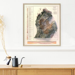 Puede incluir: Un mapa topogr&aacute;fico de Michigan de estilo vintage, impreso en papel con fondo crema. El mapa muestra las formaciones geol&oacute;gicas del estado en tonos de verde, marr&oacute;n y amarillo. El mapa est&aacute; enmarcado en un sencillo marco dorado.