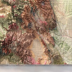 May include: A detailed, three-dimensional relief map with a vintage aesthetic. The map features a complex topography with varying shades of brown, green, and pink, suggesting mountainous regions and valleys. Grid lines overlay the map, adding a classic cartographic element.