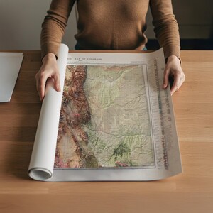 May include: A person is unrolling a vintage map of Colorado, revealing detailed topographical features in shades of green, brown, and beige. The map is printed on aged paper, with the title visible at the top. The map is on a wooden table.