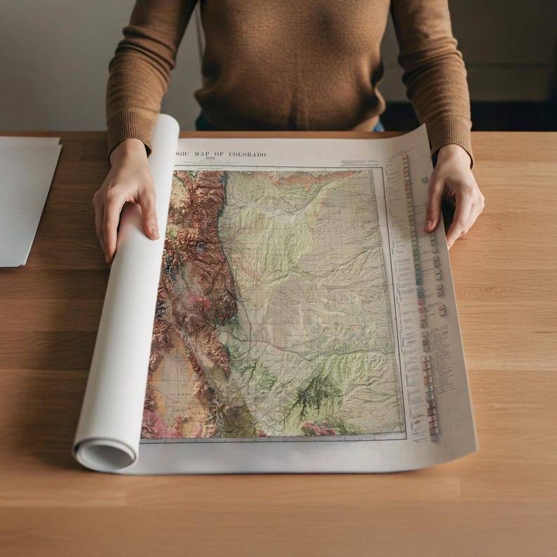 May include: A person is unrolling a vintage map of Colorado, revealing detailed topographical features in shades of green, brown, and beige. The map is printed on aged paper, with the title visible at the top. The map is on a wooden table.