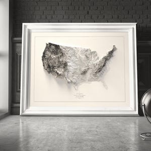 Pu&ograve; includere: Una mappa topografica in bianco e nero degli Stati Uniti d'America in una cornice bianca. La mappa &egrave; una mappa in rilievo 3D che mostra le montagne e le valli del paese. La mappa &egrave; appesa a un muro in una stanza con un muro di mattoni e una finestra.