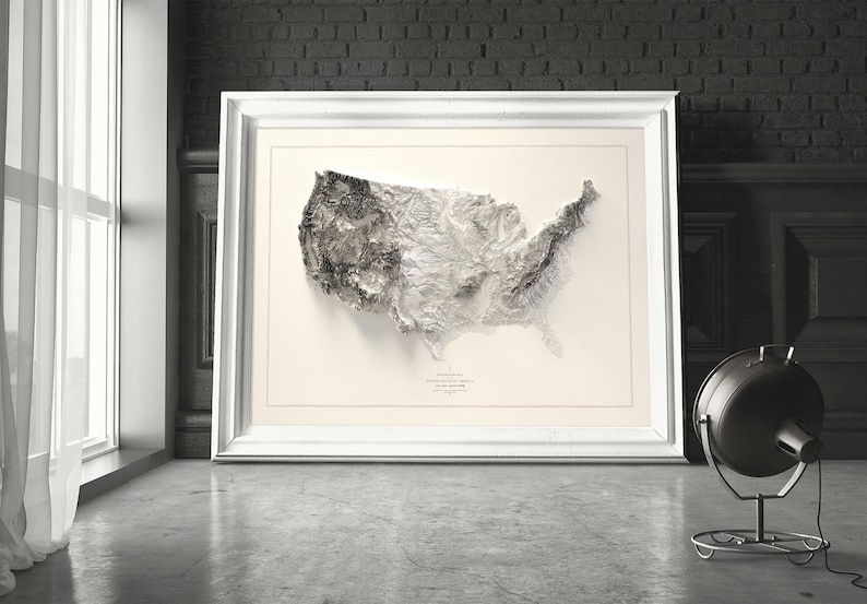 Pu&ograve; includere: Una mappa topografica in bianco e nero degli Stati Uniti d'America in una cornice bianca. La mappa &egrave; una mappa in rilievo 3D che mostra le montagne e le valli del paese. La mappa &egrave; appesa a un muro in una stanza con un muro di mattoni e una finestra.