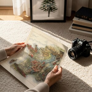 May include: A vintage map of France with raised relief, held by hands. The map features detailed topography in shades of green, brown, and red. A black camera with a lens sits nearby. The map's title is in French.