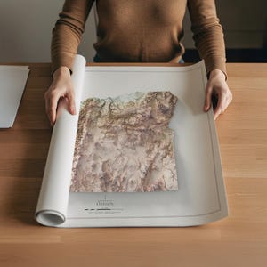 Puede incluir: Una persona sostiene una impresi&oacute;n blanca enrollada de Oreg&oacute;n. El mapa presenta un dise&ntilde;o topogr&aacute;fico 3D en tonos beige, marr&oacute;n y verde. La palabra "Oregon" est&aacute; impresa en la parte inferior del mapa.