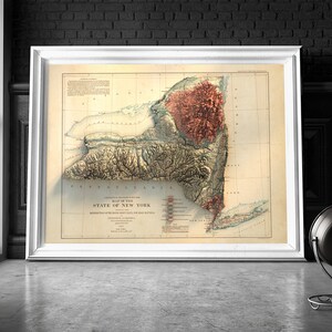 May include: A framed vintage map of New York State, featuring detailed topographical relief. The map is in a white frame, leaning against a wall. The map's colors include beige, brown, and red, with text at the bottom. A black lamp is in the foreground.