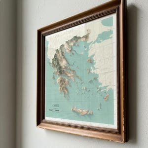 May include: A framed topographic map of Greece featuring a light teal ocean background and textured, raised-relief landmasses in shades of beige and brown. The map includes a grid overlay, the text GREECE, and a scale bar in the lower left corner.