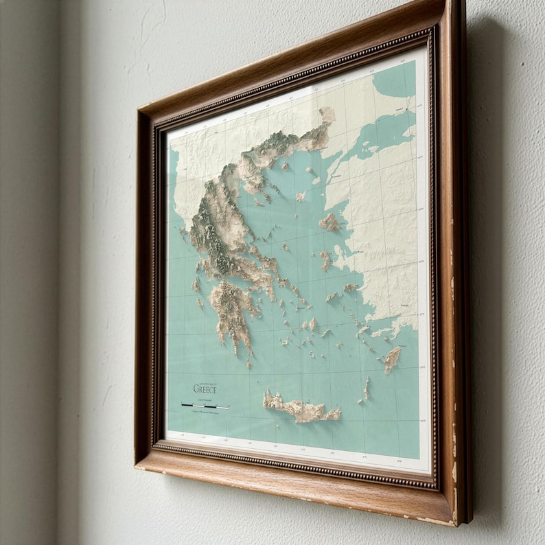 May include: A framed topographic map of Greece featuring a light teal ocean background and textured, raised-relief landmasses in shades of beige and brown. The map includes a grid overlay, the text GREECE, and a scale bar in the lower left corner.