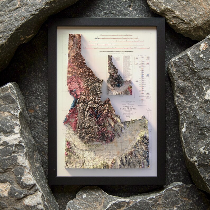 May include: Framed 3D topographic map of Idaho, featuring detailed elevation data in shades of brown, pink, and green. The map is set against a white background within a black frame, showcasing the state's geographical features.