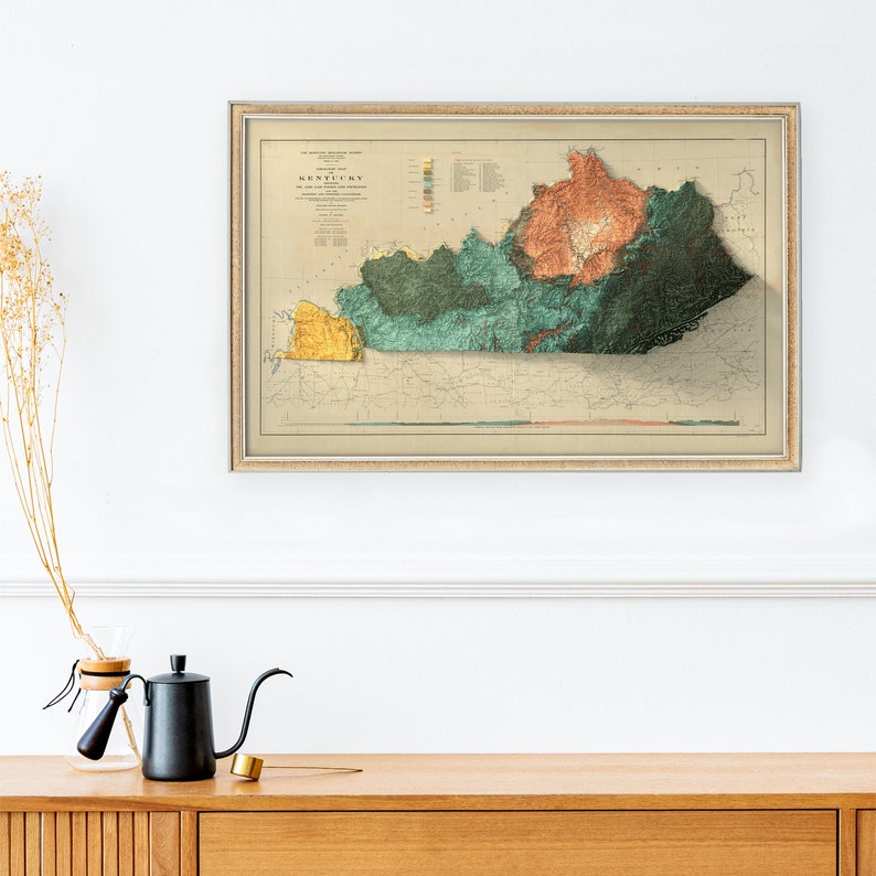May include: A vintage map of Kentucky with a 3D effect, showing the state's topography and elevation. The map is framed in a light brown frame with a cream-colored mat.