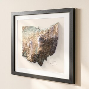 K&ouml;nnte beinhalten: Gerahmte 3D-Topografische Karte von Ohio, mit detaillierter Reliefdarstellung in Braun-, Beige- und Gr&uuml;nt&ouml;nen. Die Karte befindet sich vor einem cremefarbenen Hintergrund, gerahmt in einem schwarzen, rechteckigen Rahmen mit wei&szlig;em Passepartout.