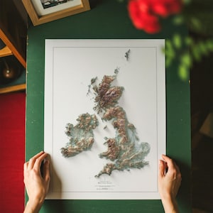 May include: A framed relief map of the British Isles, showcasing the topography with varying shades of brown and green. The map is set against a white background, with the title "British Isles" at the bottom. The frame is a light color.