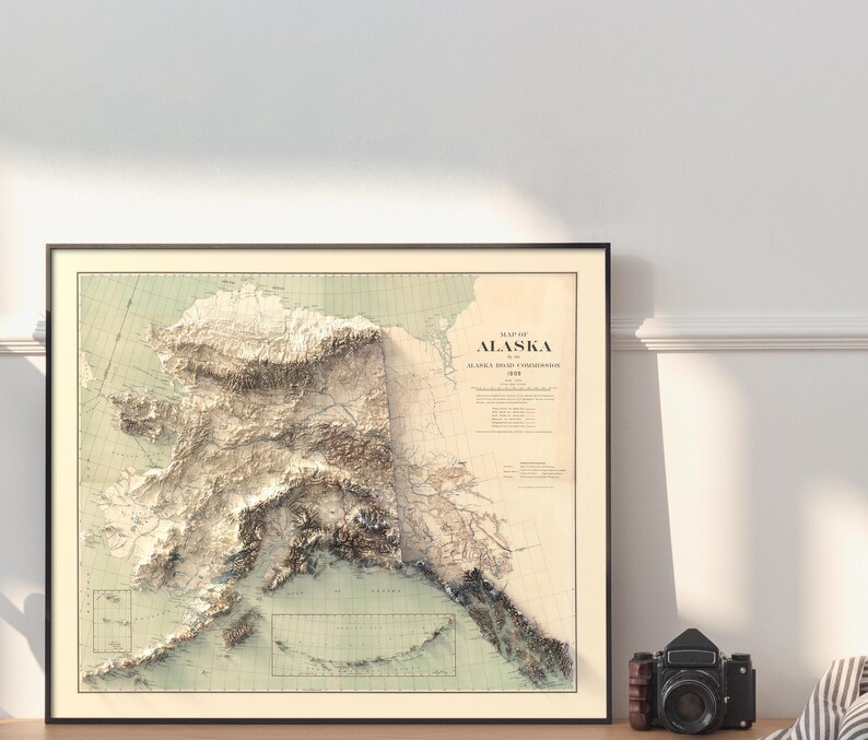 Sleek black-framed Alaska map art in a contemporary living room with neutral tones, potted plants, and minimalist shelving. The 3D elevation details of the Shelikof Strait and Kenai Fjords add bold texture to the space, perfect for urban explorers who crave a touch of Arctic wildness in their modern home or office decor.