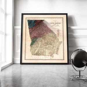 May include: A vintage map of the state of Georgia, printed in black and white with colored accents. The map is framed in a black frame and is hanging on a white wall.