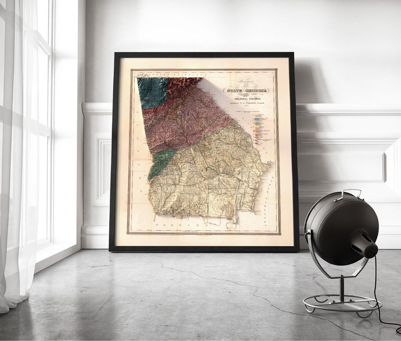 May include: A vintage map of the state of Georgia, printed in black and white with colored accents. The map is framed in a black frame and is hanging on a white wall.