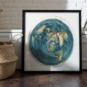 May include: A framed artwork depicting a detailed, three-dimensional rendering of Earth. The globe features shades of blue for oceans and green and tan for continents. The artwork is encased in a black frame and rests against a white wall.