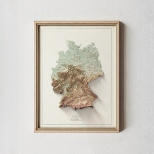 May include: A framed 3D relief map of Germany, depicting the country's terrain in brown, green, and beige hues. The map is set against a white backdrop and enclosed in a light wood-effect frame. The word "Germany" is printed at the bottom.