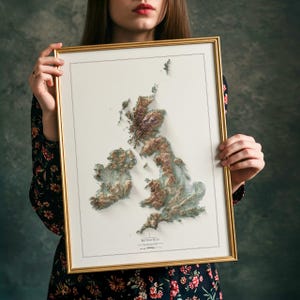 May include: Framed 3D relief map of the British Isles, featuring detailed topographical features in shades of brown and green. The map is set against a white background and framed in a gold-colored frame.