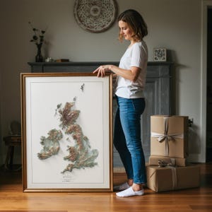 May include: A framed 3D relief map of the British Isles, featuring detailed topographical features in shades of brown, green, and white. The map is displayed in a gold-colored wooden frame. The image is of a woman holding the framed map.