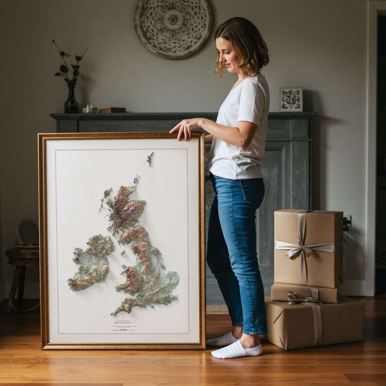 May include: A framed 3D relief map of the British Isles, featuring detailed topographical features in shades of brown, green, and white. The map is displayed in a gold-colored wooden frame. The image is of a woman holding the framed map.