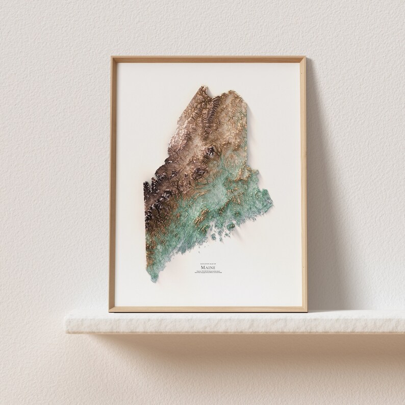 May include: A framed print of a topographic map of the state of Maine. The map is in shades of brown, green, and blue, and shows the state's mountains, rivers, and coastline.