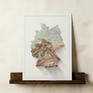 May include: A framed 3D relief map of Germany, depicting the country's topography in green, brown, and beige hues. The map is set against a white backdrop and framed in white. It rests on a dark wooden shelf, with the word "Germany" visible.
