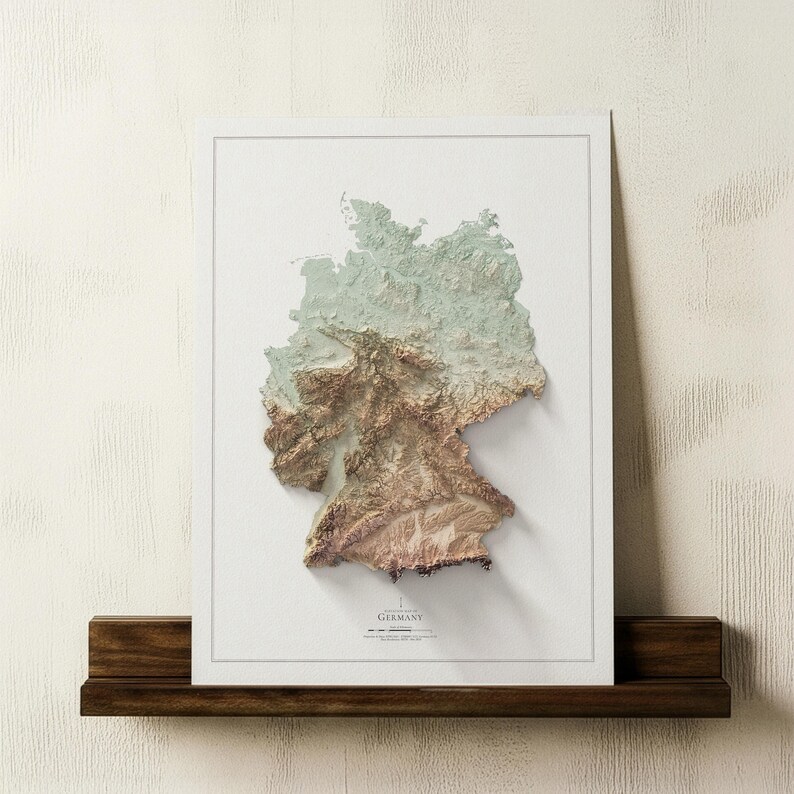 May include: A framed 3D relief map of Germany, depicting the country's topography in green, brown, and beige hues. The map is set against a white backdrop and framed in white. It rests on a dark wooden shelf, with the word "Germany" visible.