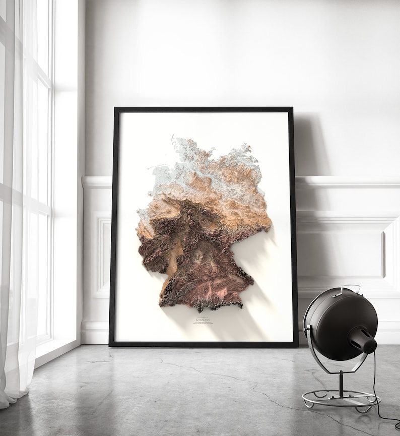 May include: A framed print of a topographic map of Germany. The map is in shades of brown, beige, and white, with a shadow cast below. The word "Germany" is printed in black at the bottom of the map.