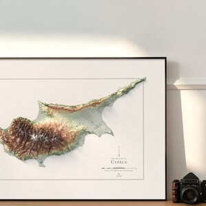 May include: A topographic map of Cyprus, showing the island's terrain in shades of green, brown, and white. The map is framed in a black frame and is hanging on a white wall.