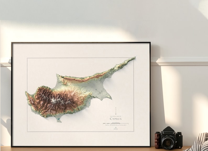 May include: A topographic map of Cyprus, showing the island's terrain in shades of green, brown, and white. The map is framed in a black frame and is hanging on a white wall.