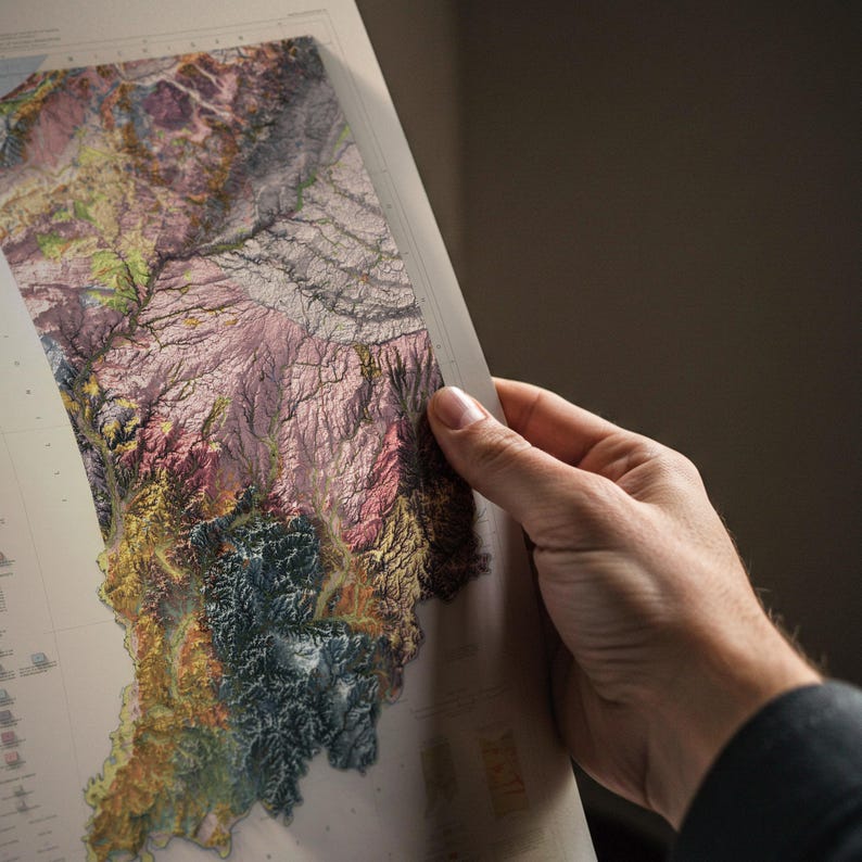 May include: A detailed topographic map with a vibrant colour palette including pink, green, yellow, and blue. The map, held by a person, highlights the intricate details of the terrain. It appears to be a geographical representation of a specific region.