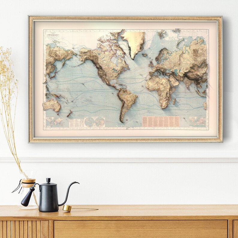 May include: A framed world map print featuring a raised relief style with golden-toned mountain ranges and landmasses set against a pale blue ocean background. The map includes detailed topographical shading, thin blue latitude lines, and text.