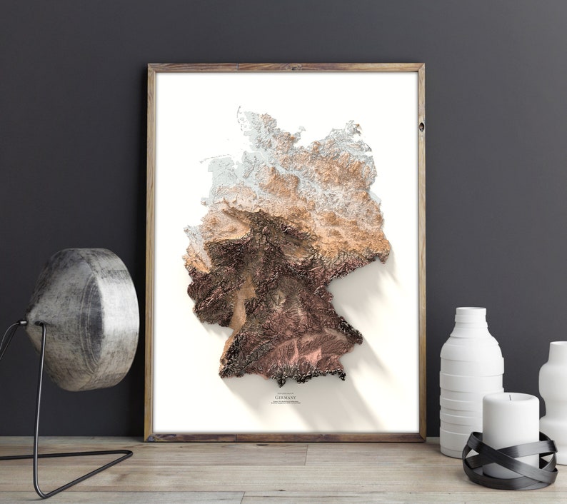 May include: A framed print of a 3D topographic map of Germany. The map is in shades of brown, beige, and white, with a shadow effect. The word "GERMANY" is printed at the bottom of the map.
