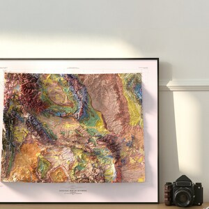 Puede incluir: Un mapa geol&oacute;gico enmarcado de Wyoming con un dise&ntilde;o colorido, texturizado y con un aspecto 3D. El mapa muestra las diferentes formaciones rocosas y caracter&iacute;sticas geol&oacute;gicas del estado.
