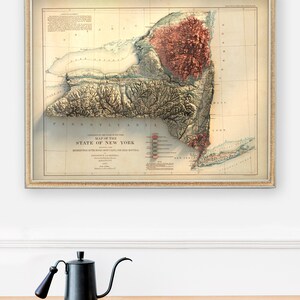 May include: Framed vintage map of New York State with a 3D relief effect, showcasing the state's topography. The map is in a gold frame and hangs above a wooden cabinet. A black gooseneck kettle with a wooden handle sits on the cabinet.