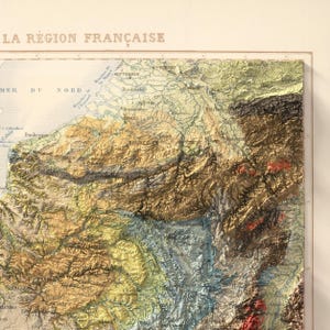 May include: A detailed, vintage relief map of a region in France, featuring textured terrain in shades of brown, green, and yellow. The map includes place names and is labeled "LA R&Eacute;GION FRAN&Ccedil;AISE" at the top. The map is framed.