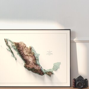 May include: Framed 3D topographic map of Mexico, featuring detailed relief in shades of brown, green, and blue. The map is set against a cream background within a black frame. A vintage camera sits nearby.