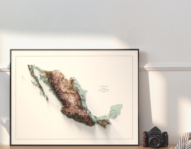 May include: Framed 3D topographic map of Mexico, featuring detailed relief in shades of brown, green, and blue. The map is set against a cream background within a black frame. A vintage camera sits nearby.