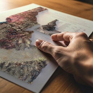 May include: A detailed 3D topographical map with a hand holding a clear overlay. The map features various colors, including shades of brown, red, and green, representing different elevations. The map is on a white background.