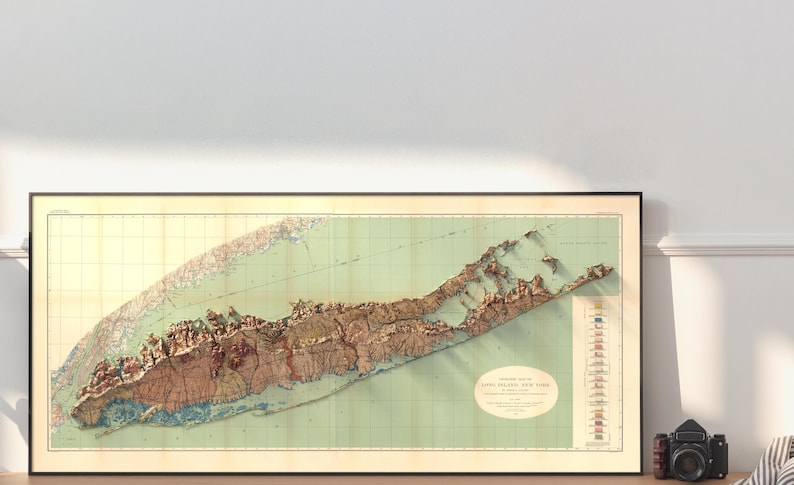 May include: A vintage, detailed relief map of Long Island, New York, printed on paper with a green background and brown relief features. The map is framed in a black frame and includes a legend with colored flags.