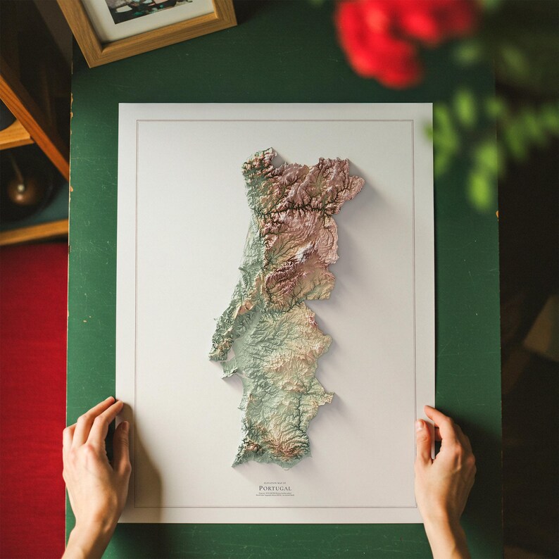 May include: A framed 3D relief map of Portugal, showcasing the country's topography in shades of green, brown, and beige. The map is set against a white background, with the word "PORTUGAL" printed at the bottom. The frame is a light color.