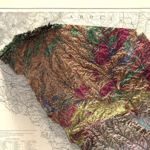 Pode incluir: Um mapa de relevo vintage da Carolina do Sul, mostrando a topografia do estado com diferentes cores que representam diferentes eleva&ccedil;&otilde;es. O mapa &eacute; impresso em papel e tem uma borda detalhada com as palavras "Carolina do Norte" no topo.