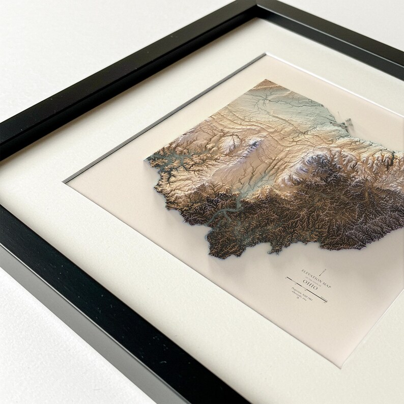 K&ouml;nnte beinhalten: Gerahmte 3D-H&ouml;henkarte von Ohio, die die Topographie des Staates in Braun-, Beige- und Blaut&ouml;nen zeigt. Die Karte ist in einem wei&szlig;en Passepartout und einem schwarzen Rahmen eingefasst und bietet eine moderne, k&uuml;nstlerische Darstellung der Landschaft.
