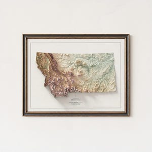 May include: A framed 3D topographical map of Montana, USA, in shades of brown, green, and beige. The map is set within a dark brown frame with a white border. The word "Montana" is printed at the bottom.