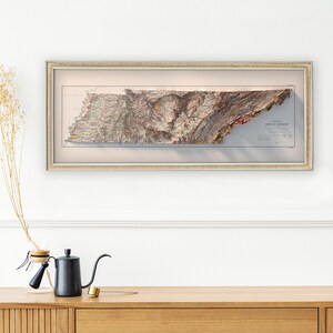 May include: A framed, vintage-style, topographic map of the state of Tennessee. The map is in shades of brown, green, and blue, and shows the state's mountains and rivers. The map is framed in a light brown frame.