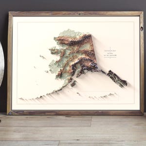 May include: A framed 3D relief map of Alaska, showcasing the state's topography in shades of brown, green, and white. The map is set against a white background within a wooden frame. The text "Elevation Map Alaska" is visible.