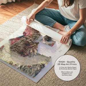 May include: A detailed 2D map art print of Idaho, featuring topographical details in shades of brown, red, and green. The map is being unrolled, revealing the state's contours. The text on the print reads "STATE OF IDAHO". The print is on matte paper.