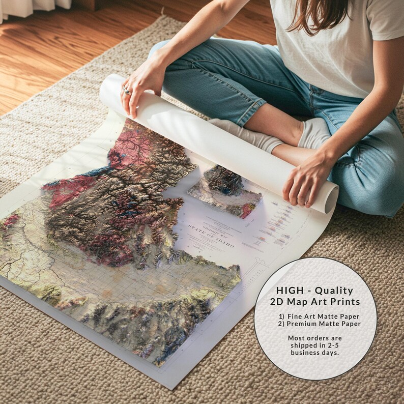 May include: A detailed 2D map art print of Idaho, featuring topographical details in shades of brown, red, and green. The map is being unrolled, revealing the state's contours. The text on the print reads "STATE OF IDAHO". The print is on matte paper.